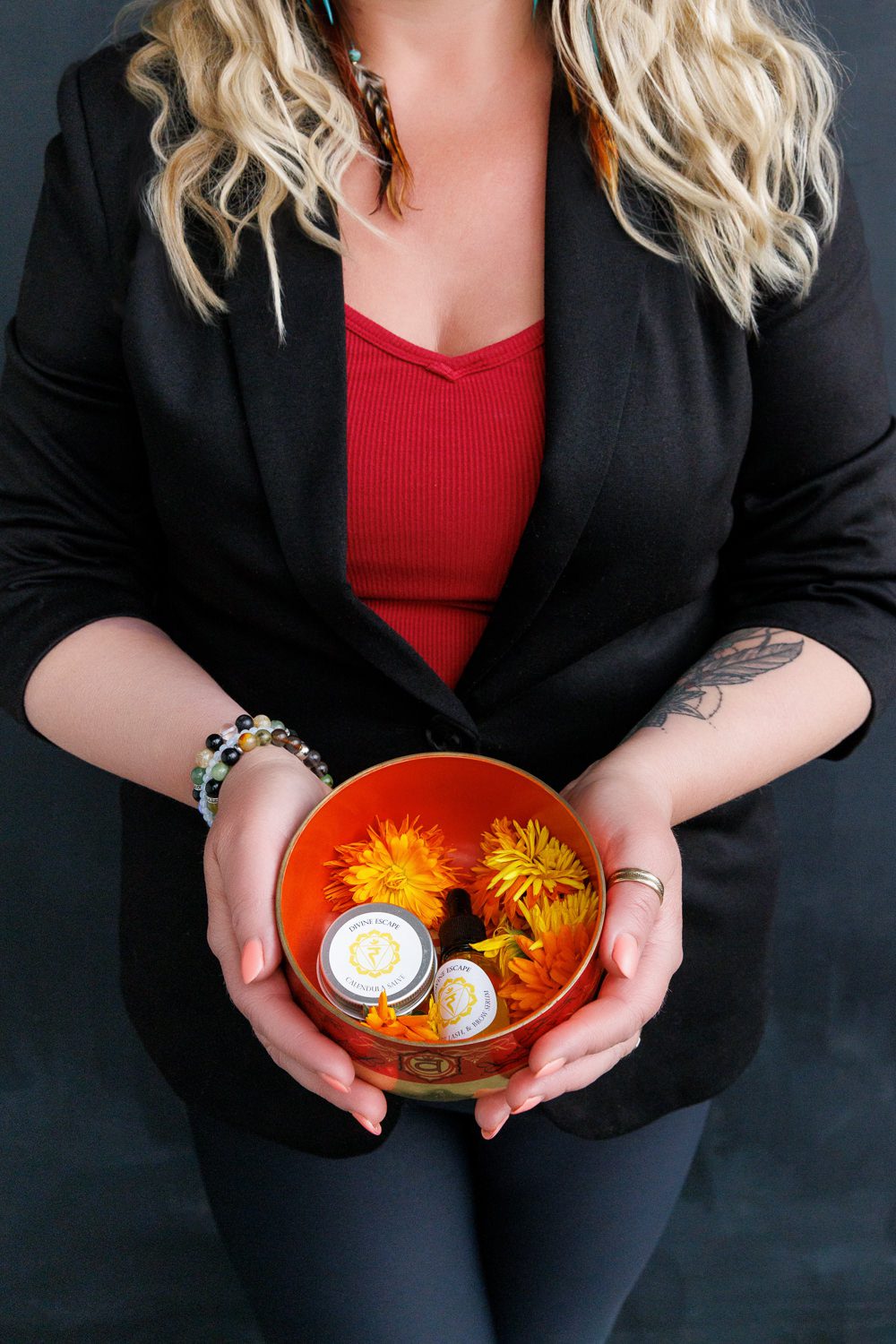 Close-up of a woman in a black blazer and red top holding a red singing bowl filled with yellow flowers and small natural skincare products by Divine Escape during a wellness branding mini session in Calgary.